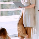 Kids Corner - Adult female wearing casual clothes standing in corner of room and using laptop while spending time with daughter in daytime