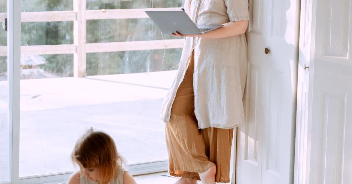 Kids Corner - Adult female wearing casual clothes standing in corner of room and using laptop while spending time with daughter in daytime